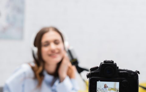 Digital camera on tripod near blurred brunette broadcaster in headphones using laptop near microphone during stream in podcast studio